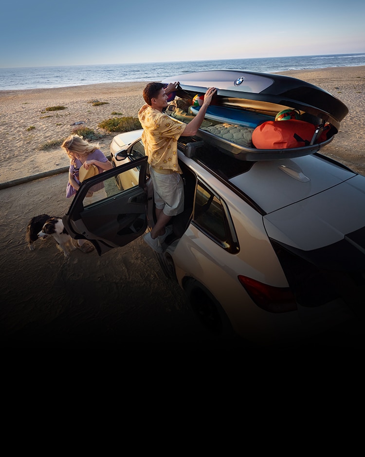 A man, standing on the door sill, reaches into a BMW Roof Box atop a white BMW X3 that is parked on the beach facing the ocean. A woman stands behind the man tending to a small brown and white dog.