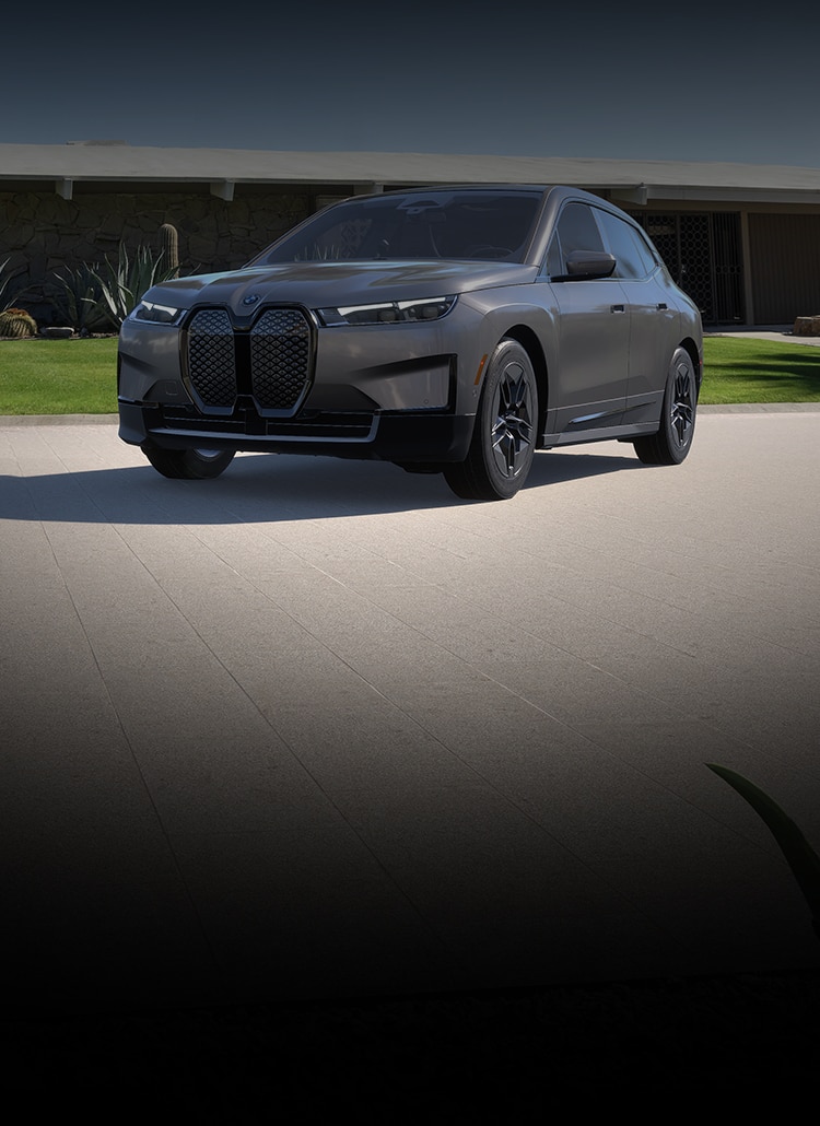 The 2025 BMW iX xDrive50 parked in front of a modern home with a dramatic shadow of the vehicle on the driveway.
