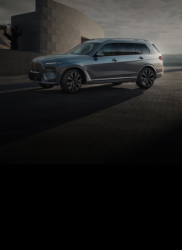 The 2025 BMW X7 xDrive40i parked in front of a modern stone building with mountains in the background.
