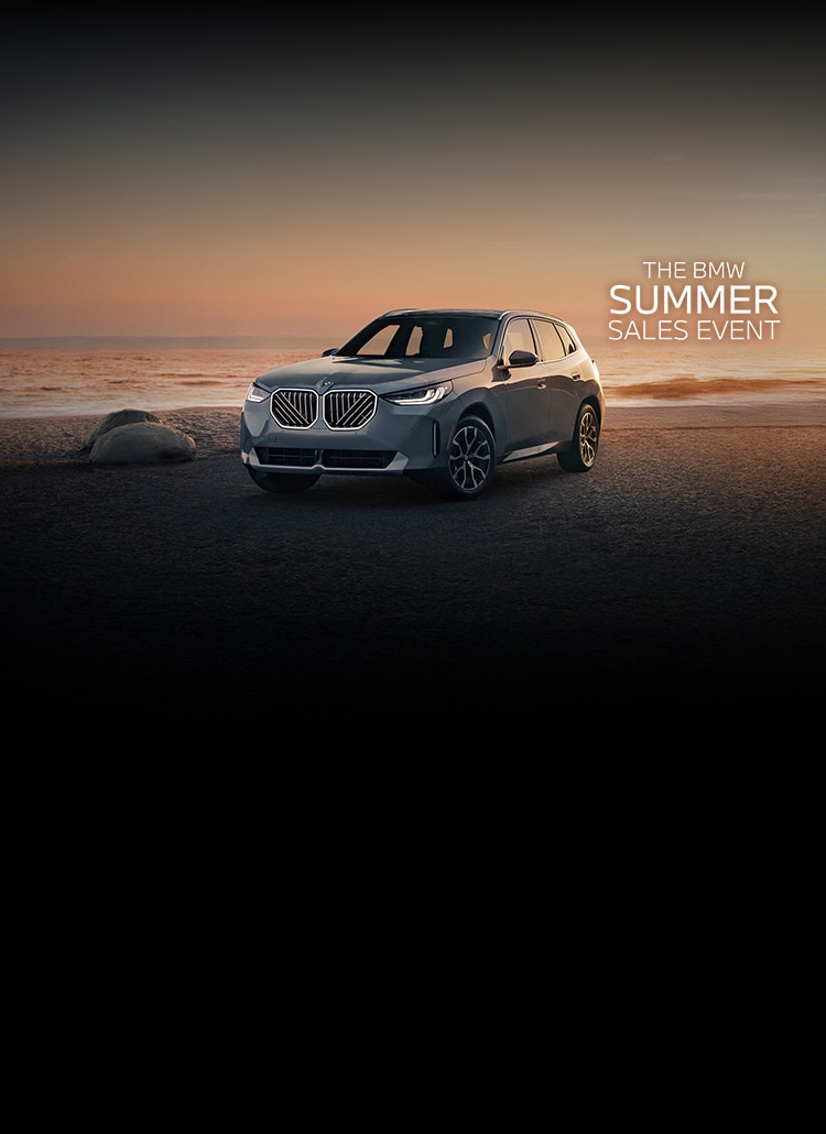 The 2025 X3 30 xDrive SUV parked at the beach at sunset with headlights aglow.