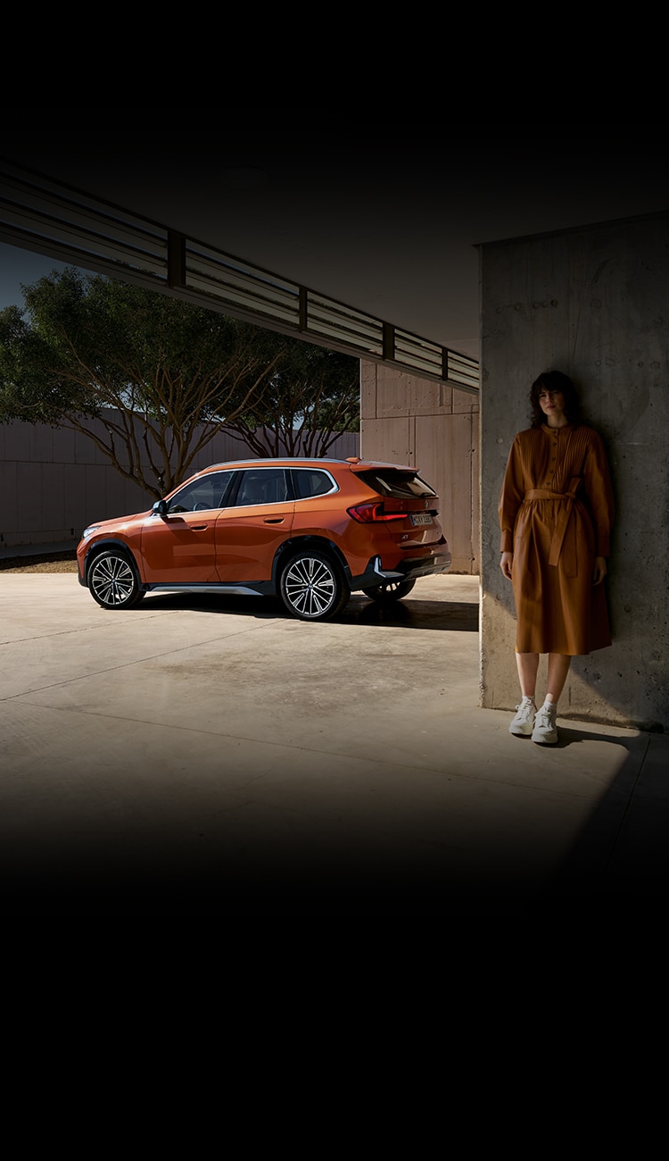 A woman stands near the 2023 BMW X1 SAV in Utah Orange Metallic parked on a driveway