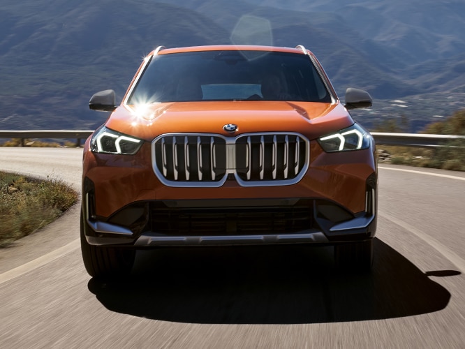Heroic front-on shot of the 2023 BMW X1 compact SAV with Full LED headlights illuminated while driving down a curvy road