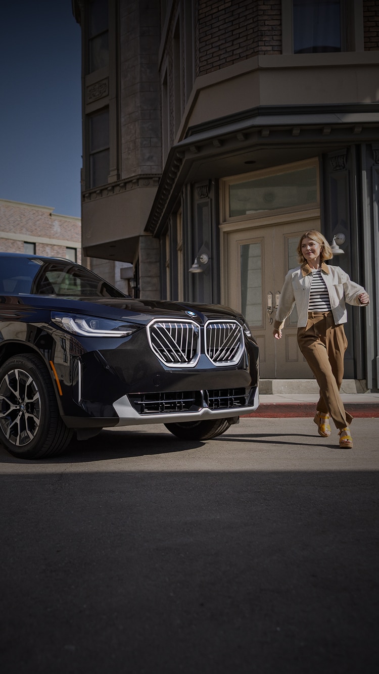A woman walks around the front of her BMW X3 30 xDrive after she parks it in an urban environment