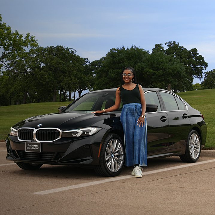A girl leans on a BMW 3 Series in a parking lot next to a large park.