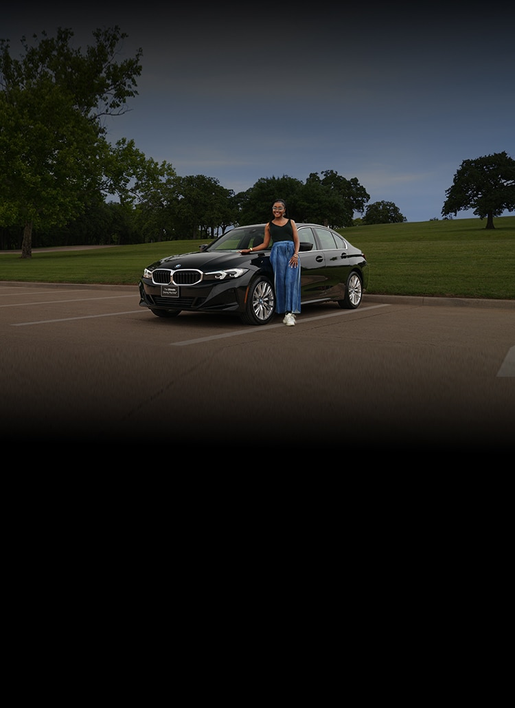 A girl leans on a BMW 3 Series in a parking lot next to a large park 