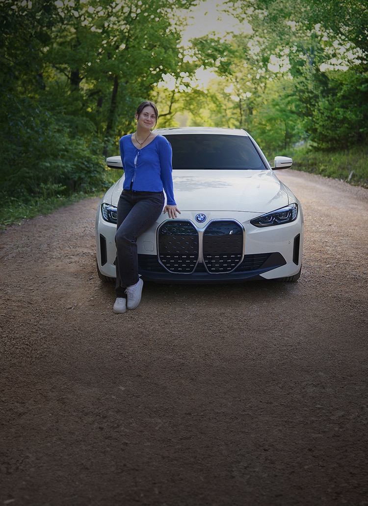 A smiling young woman leans on the front hood of a BMW i4 on a gravel road
