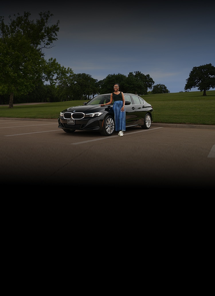 A girl leans on a BMW 3 Series in a parking lot next to a large park 
