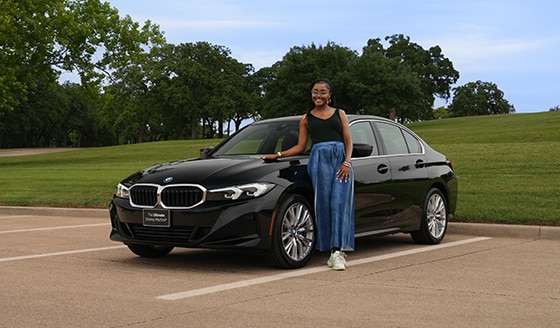 A girl leans on a BMW 3 Series in a parking lot next to a large park 