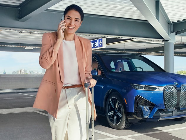 Image of a woman walking away from her parked BMW with a rolling suitcase in one hand, smartphone in the other