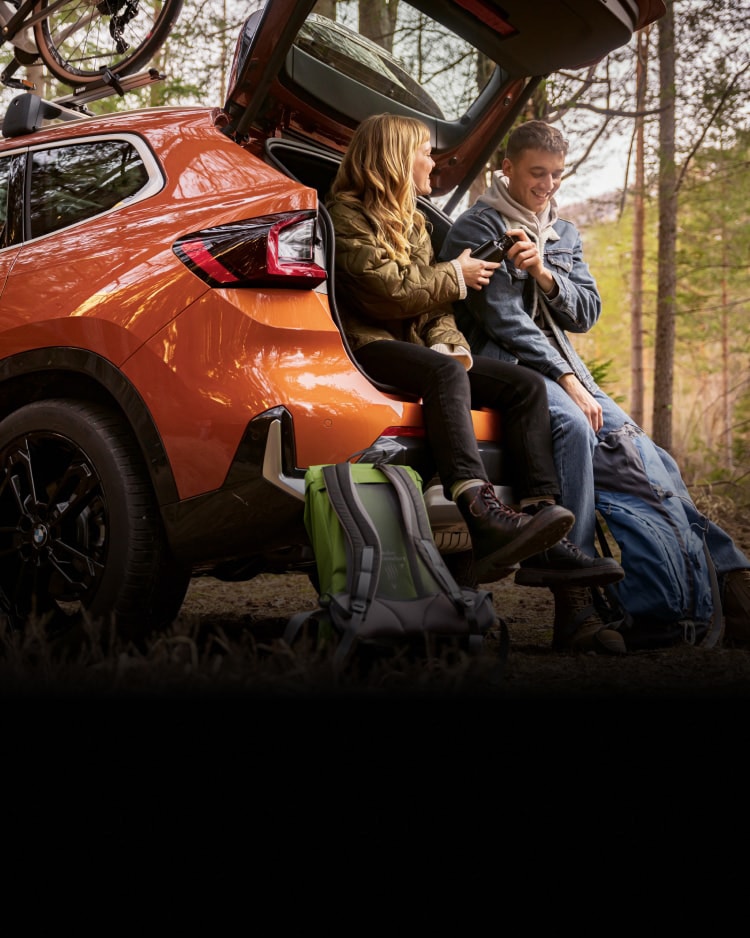 Two young people sitting in the open trunk of their BMW in a forested area.