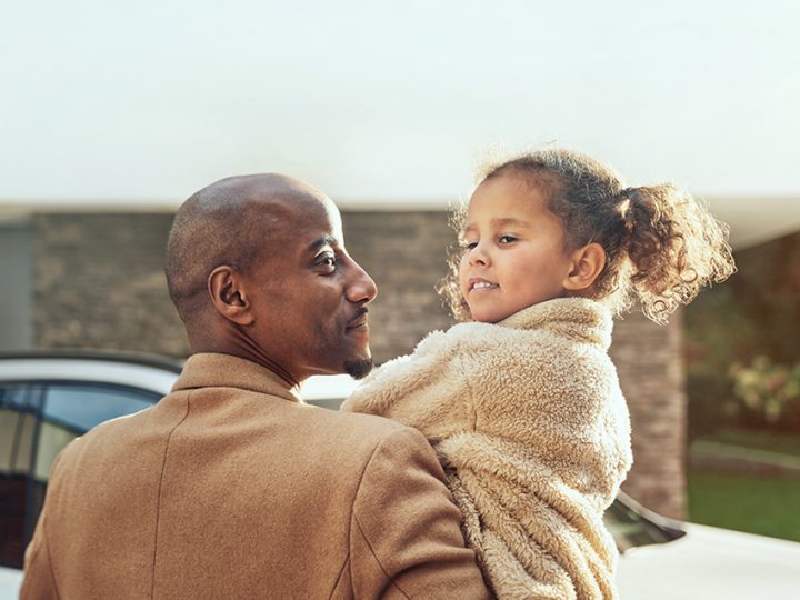 An adoring father looks over at his daughter while carrying her to a parked BMW