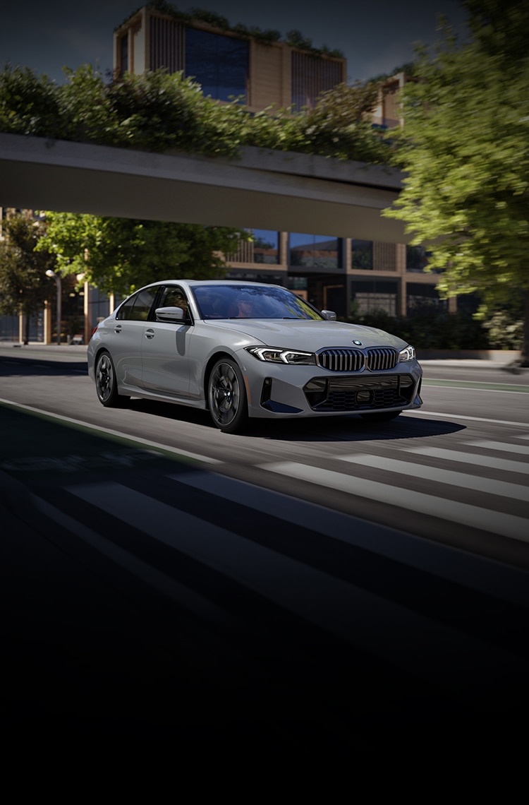 A casually-dressed man stands on a driveway in front of a parked BMW