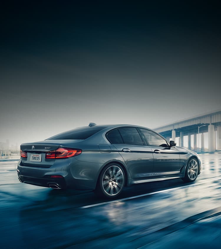 BMW 5 Series Sedan approaching a bridge on the road with city in the background