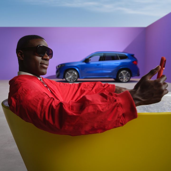 A BMW driver in a vibrant red jacket uses the My BMW app on his smartphone while his his BMW is parked in the background.