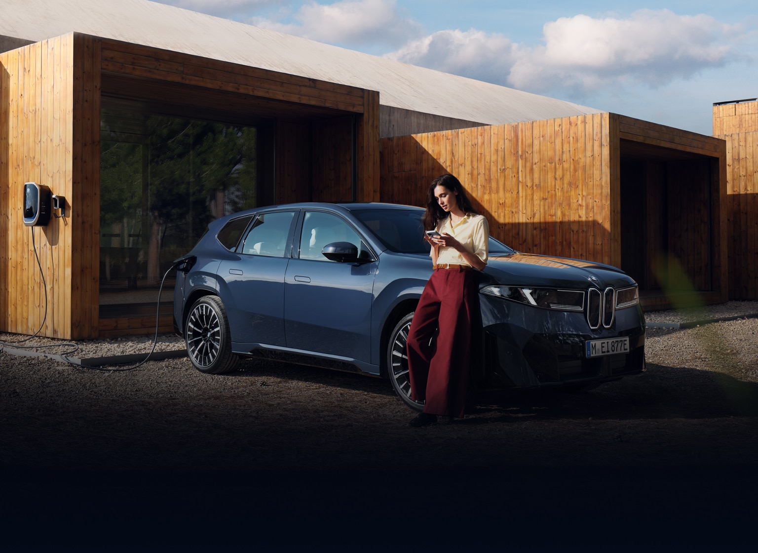 A BMW driver leans against her charging BMW while using the My BMW app on her smartphone.