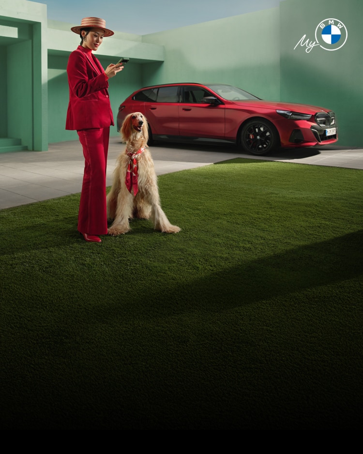 A BMW driver in a jaunty red pantsuit and striped straw hat uses her My BMW app while her large dog relaxes next to her.