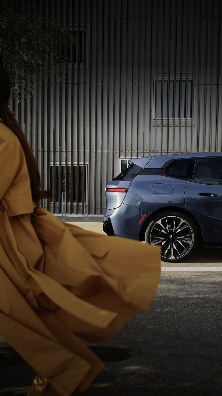 A woman walks out of the left side of the shot as a BMW iX is parked behind her.