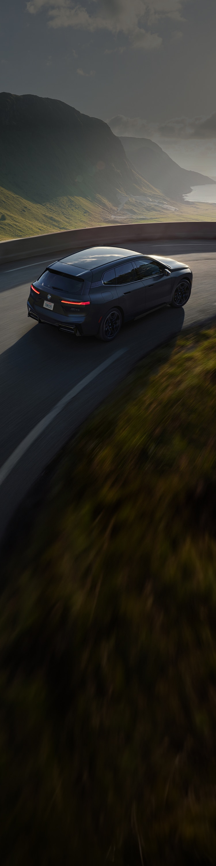 A BMW iX taking a curve on an elevated highway through a lush seaside setting.