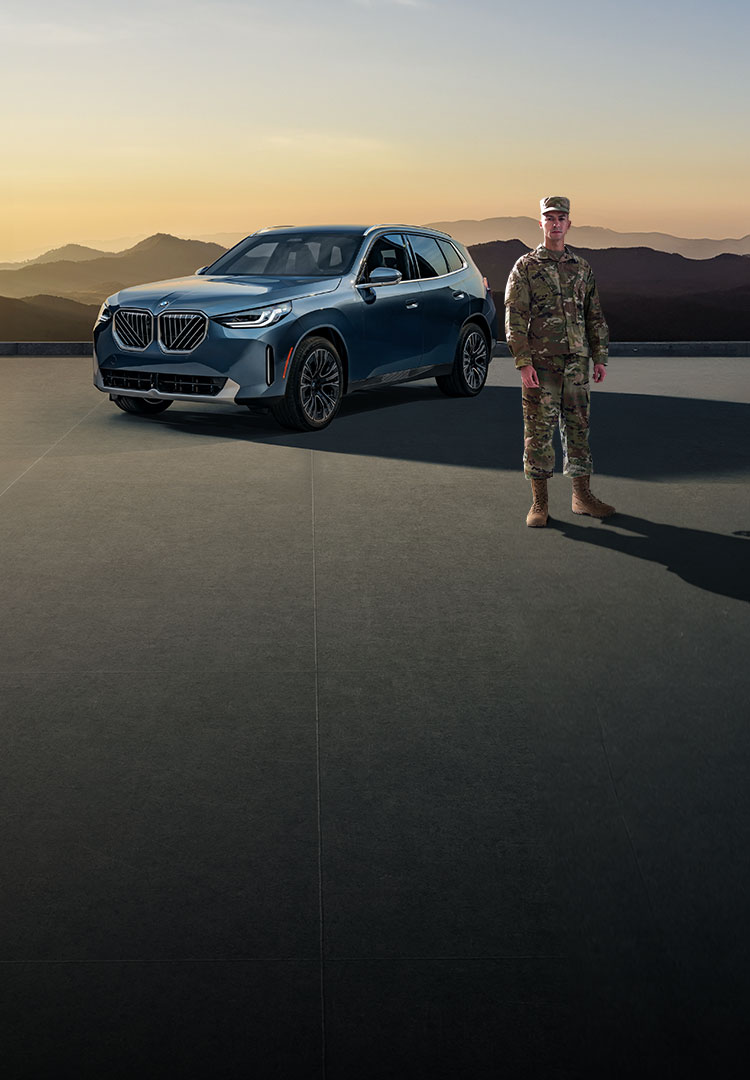 A man in military fatigues stands next to a BMW X3 with a shaded forest landscape in the background