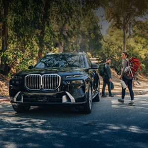 A woman and man walk away from their parked BMW X7 xDrive40i as they head off for a hike on a wooded path