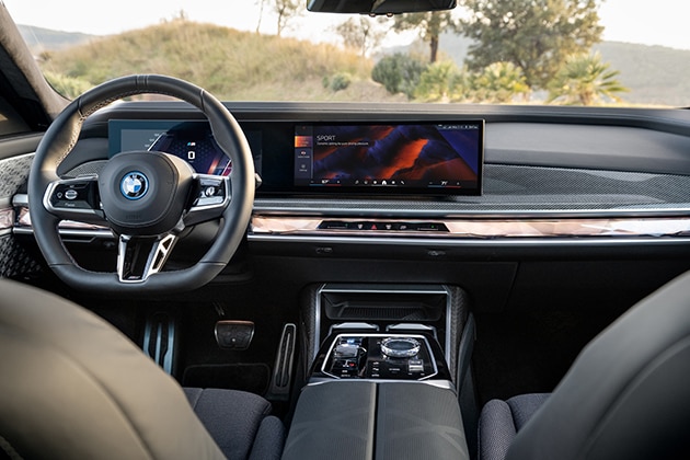 A shot front from the back seat showing the steering wheel, control console, curved display, and front seats of the BMW i7